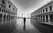 Colloquium image (C) Matjaz Licer : Black and white picture of the San Marco square in Venice during an Aqua Alta event. In the center of the picture, someone stands feet in the seawater covering the square. He/she turns the back to the photographer while presumably taking a photo of the Laguna.
