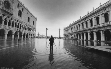 Colloquium image (C) Matjaz Licer : Black and white picture of the San Marco square in Venice during an Aqua Alta event. In the center of the picture, someone stands feet in the seawater covering the square. He/she turns the back to the photographer while presumably taking a photo of the Laguna.